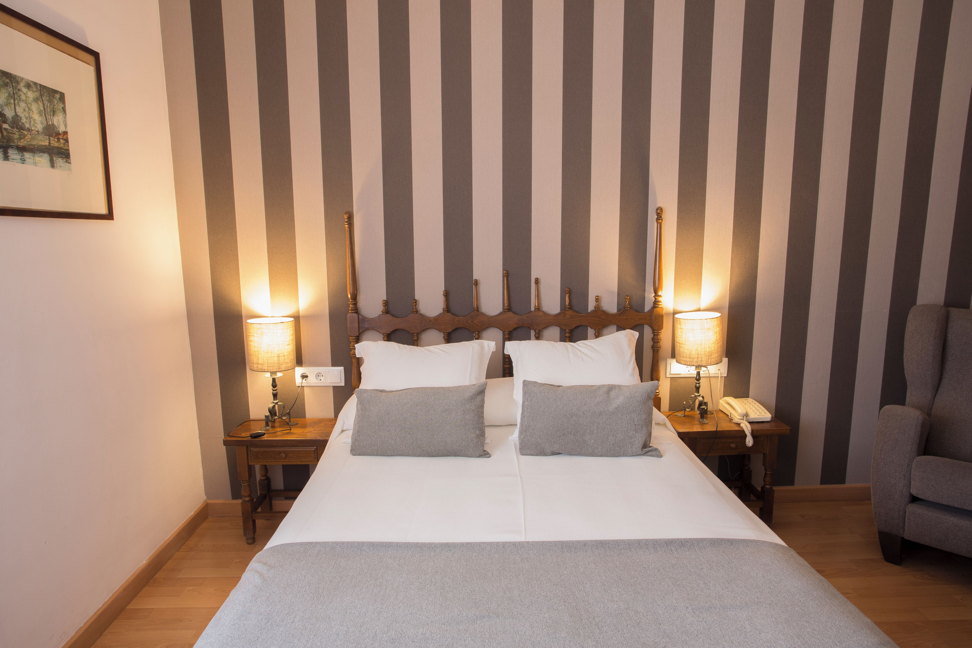 A hotel room with a large bed, two bedside tables, a chair, and a striped wall.