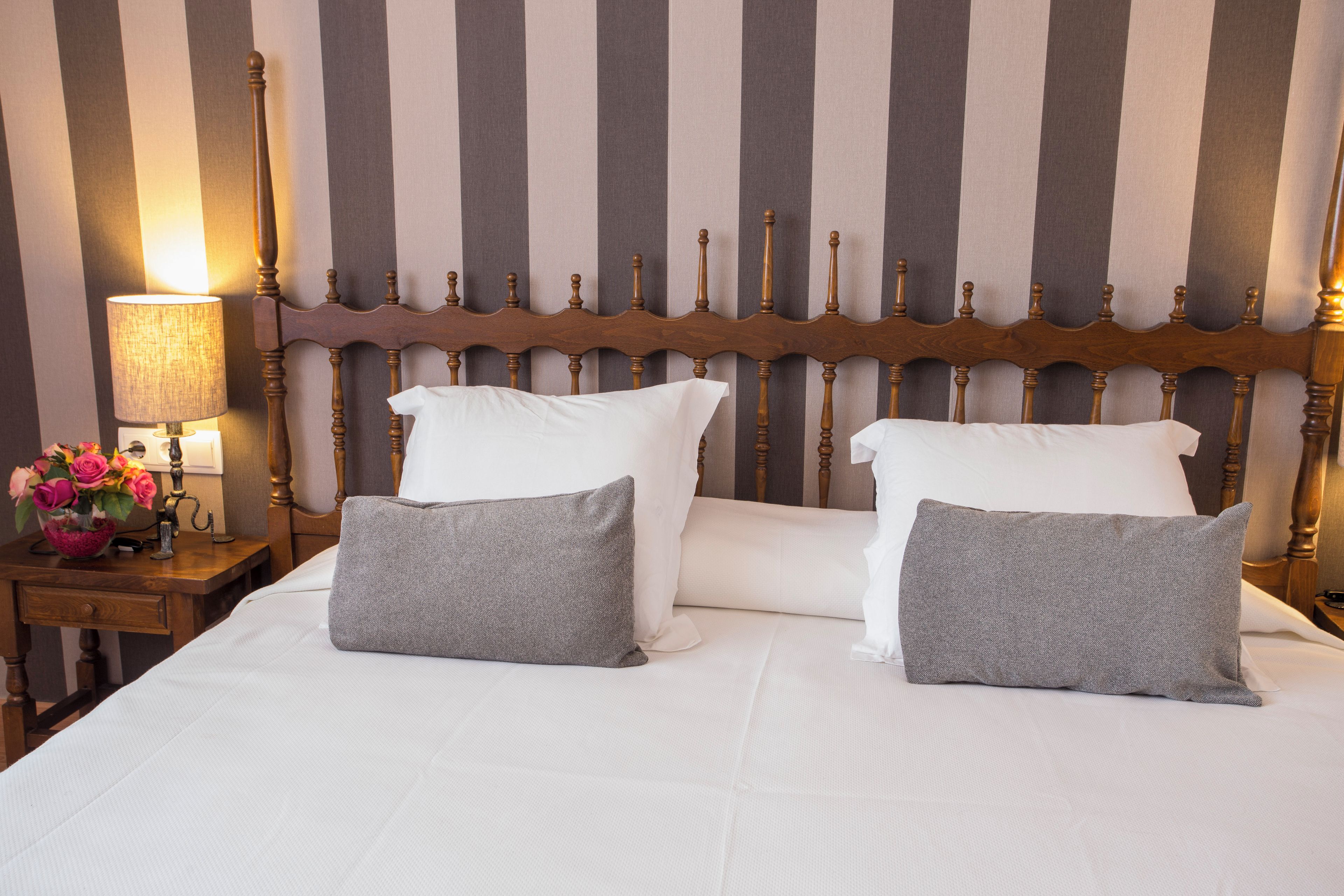 Uma cama bem arrumada com almofadas brancas e cinzentas, uma cabeceira de madeira e uma mesa de cabeceira com abajur e flores.