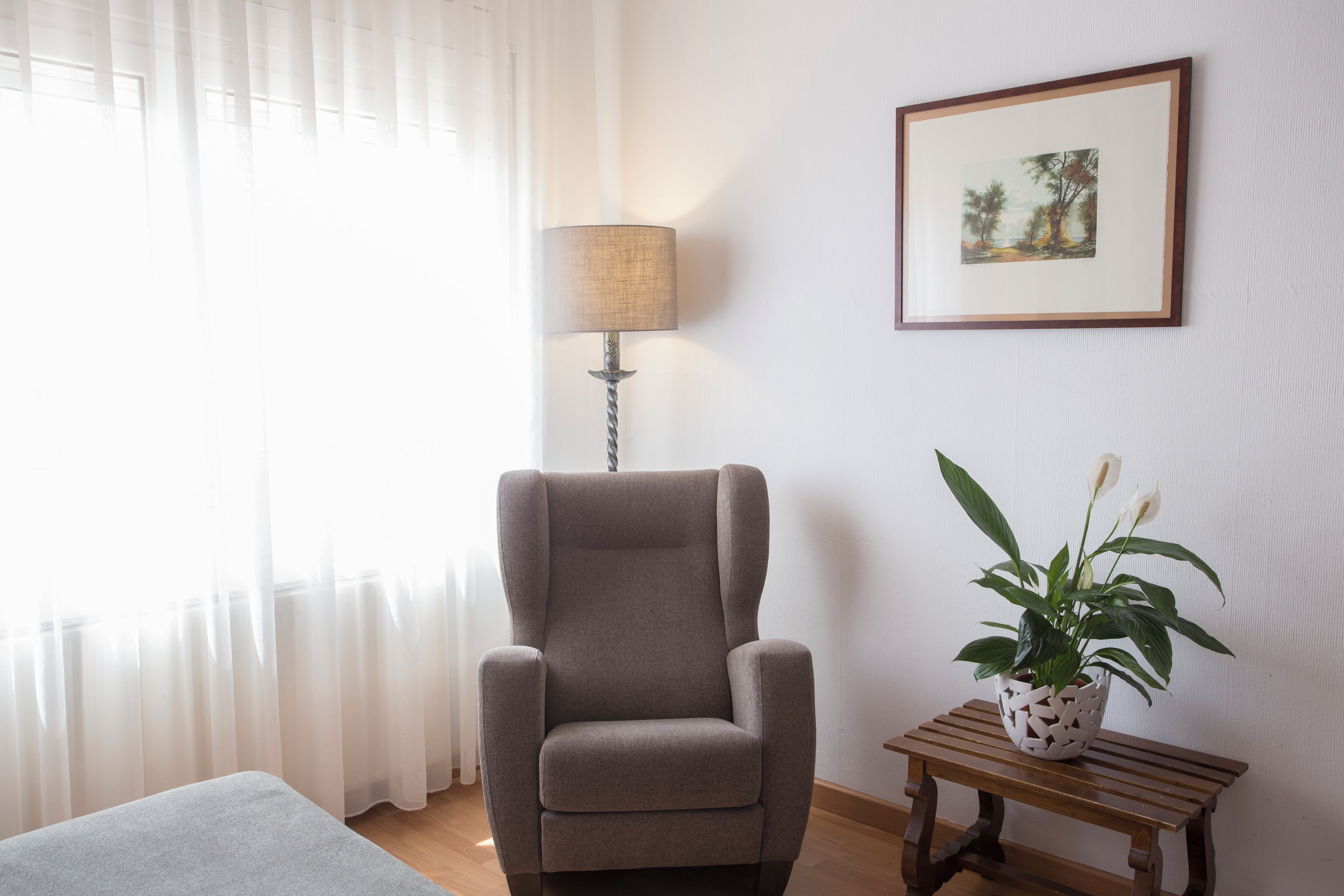 Um quarto com uma poltrona cinzenta, uma luminária, uma mesinha com uma planta e um quadro na parede.
