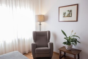 Ett rum med en grå fåtölj, en lampa, ett litet bord med en växt och en inramad tavla på väggen.