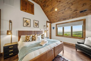 Um quarto com uma cama grande, teto de madeira, obra de arte emoldurada, mesa de cabeceira, abajur, janela com vista e um sofá.