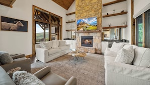 A spacious living room with a stone fireplace, a large TV, and a dining area with wooden beams and a stone wall. - Breezeway Lodge at Eagles Nest - hot tub, huge views, all Suite style rooms, close to tennis (Banner Elk)