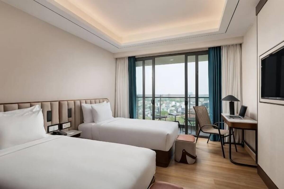 A hotel room with two beds, a desk, a chair, and a large window with a city view.