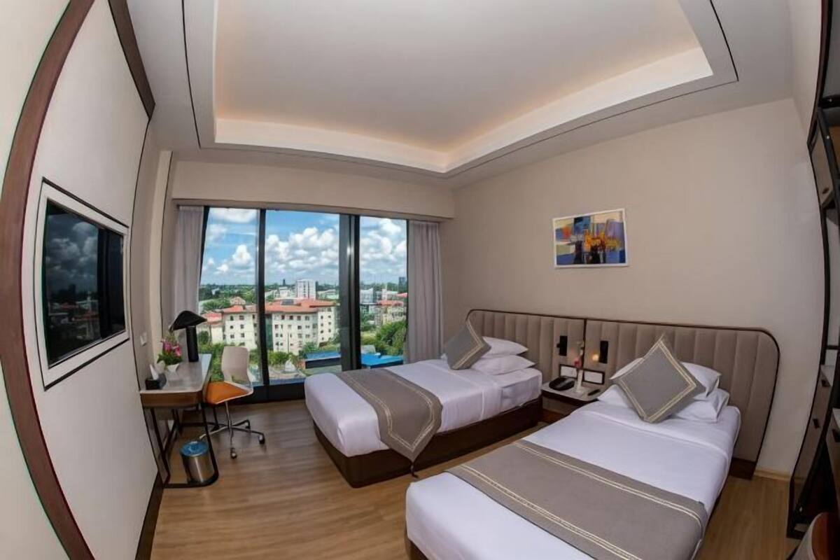 A hotel room with two beds, a desk, a chair, and a large window with a city view.