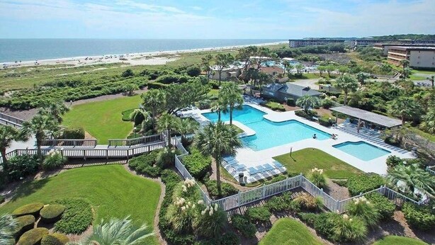 Vista aérea de un resort con una piscina central, rodeado de exuberante vegetación y una playa al fondo.