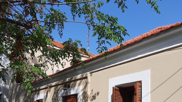 Ein Gebäude mit einer beigefarbenen Fassade, weißem Zierdekor und einem roten Ziegeldach. Es gibt drei Fenster mit Holzläden, eines davon ist geöffnet. Das Gebäude hat eine Tür und ein kleines Fenster mit einer Laterne darüber.