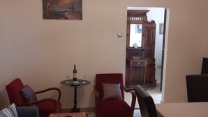 Ein Wohnzimmer mit einer Couch, Sesseln, einem Couchtisch und einer Chandelüster. An der Wand hängt ein Gemälde und eine Tür führt in eine Küche.