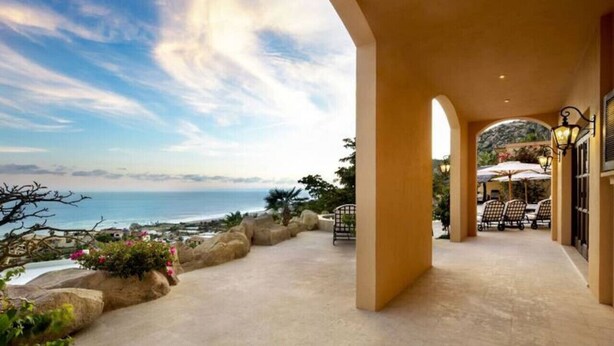 A covered outdoor area with a view of the ocean, featuring a seating area with tables and chairs, and a pathway leading to a building entrance. - Pedregal Luca incredible Views with onsite staff (Cabo San Lucas)