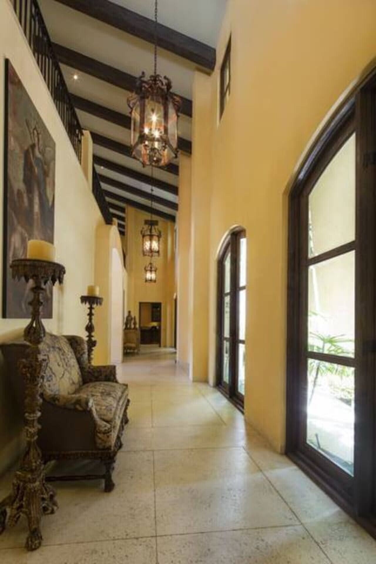 A spacious hallway with a high ceiling, ornate chandeliers, a large window, and a grand staircase.