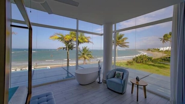 Habitación moderna con un ventanal grande con vista a la playa, una bañera y un área de descanso.