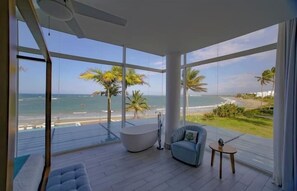 A modern room with a large window overlooking a beach, a bathtub, and a seating area.