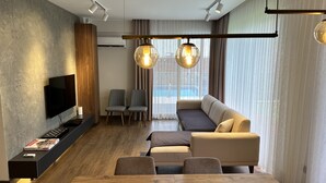 A modern living room with a flat-screen TV, a sofa, a dining table, and a large window with curtains. - DrVilla Suits (Serik)