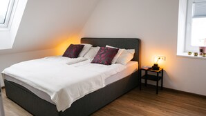 A neatly made bed with white bedding and decorative pillows, a bedside table with a lamp, and a window with blinds.