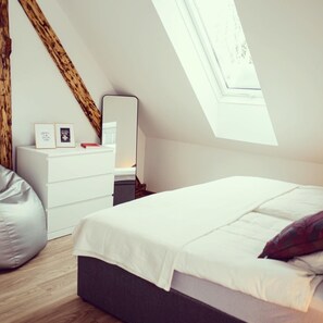 A modern bedroom with a sloped ceiling, a skylight, a white bed, a bedside table with a mirror, and a white dresser with framed pictures.