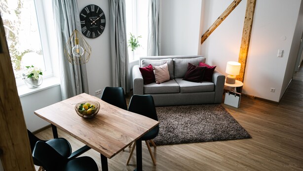 A modern living room with a wooden dining table, black chairs, a grey sofa with cushions, a black clock, and a lamp on a side table.