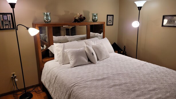 A neatly made bed with white bedding and pillows, a wooden headboard with a mirror, and two floor lamps on either side. - Stylish two bedroom Condo (Milwaukee)