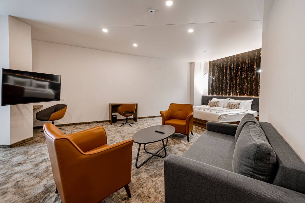A modern hotel room with a flat-screen TV, a sofa, two armchairs, a small round table, and a bed with a headboard.