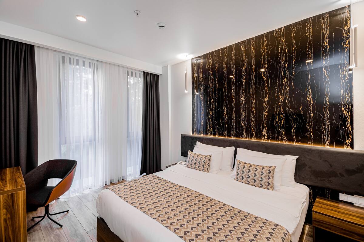 A modern hotel room with a large bed, a wooden nightstand, a chair, and a large window with sheer curtains.