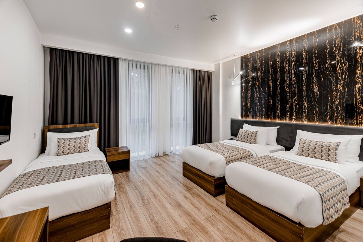 A hotel room with two beds, a large window with curtains, a wall-mounted TV, and a decorative wall panel.