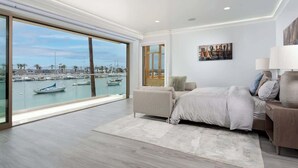 A spacious bedroom with a large window overlooking a marina, a bed, a nightstand, and a painting on the wall.