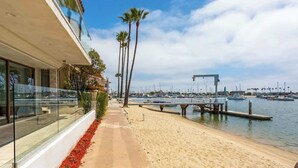 A beachfront property with a sandy walkway, palm trees, and a marina in the background.
