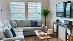 Ein modernes Wohnzimmer mit einem grauen Ecksofa, einem Holztisch, einem Fernseher auf einem Standfuß und einem großen Fenster mit Jalousien.