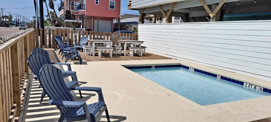 A pool area with lounge chairs and tables, surrounded by a wooden deck and a white fence.