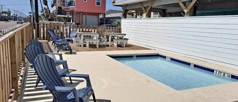 A pool area with lounge chairs and tables, surrounded by a wooden deck and a white fence.