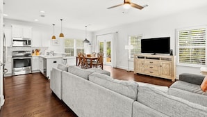 A modern kitchen with white cabinets, a dining area with a table and chairs, a gray sofa, and a television in a living room. - Amazing Inlet Beach, Shopping and Dining: Blue Mar + Free Attraction Tickets! (Inlet Beach)