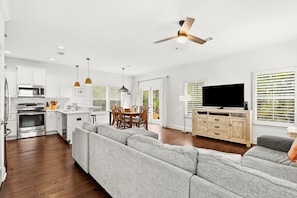 A modern kitchen with white cabinets, a dining area with a table and chairs, a gray sofa, and a television in a living room. - Amazing Inlet Beach, Shopping and Dining: Blue Mar + Free Attraction Tickets! (Inlet Beach)