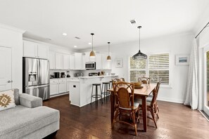 A modern kitchen with white cabinets, a stainless steel refrigerator, a dining area with a wooden table and chairs, and a large window with blinds. - Amazing Inlet Beach, Shopping and Dining: Blue Mar + Free Attraction Tickets! (Inlet Beach)