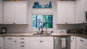 A kitchen with white cabinets, a granite countertop, a window with a view of greenery, and a 'Beach' sign above the window. - Updated near beach home, outdoor paradise, sleeps 6! (Isle of Palms)