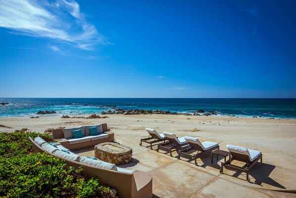 A beachside lounge area with a stone fire pit, lounge chairs, and a sofa with cushions.