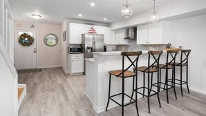A modern kitchen with white cabinets, a stainless steel refrigerator, a bar area with stools, and a decorative wreath on the door. - Large Ocean City Waterfront Townhome w/ Boat Slip (Ocean City)