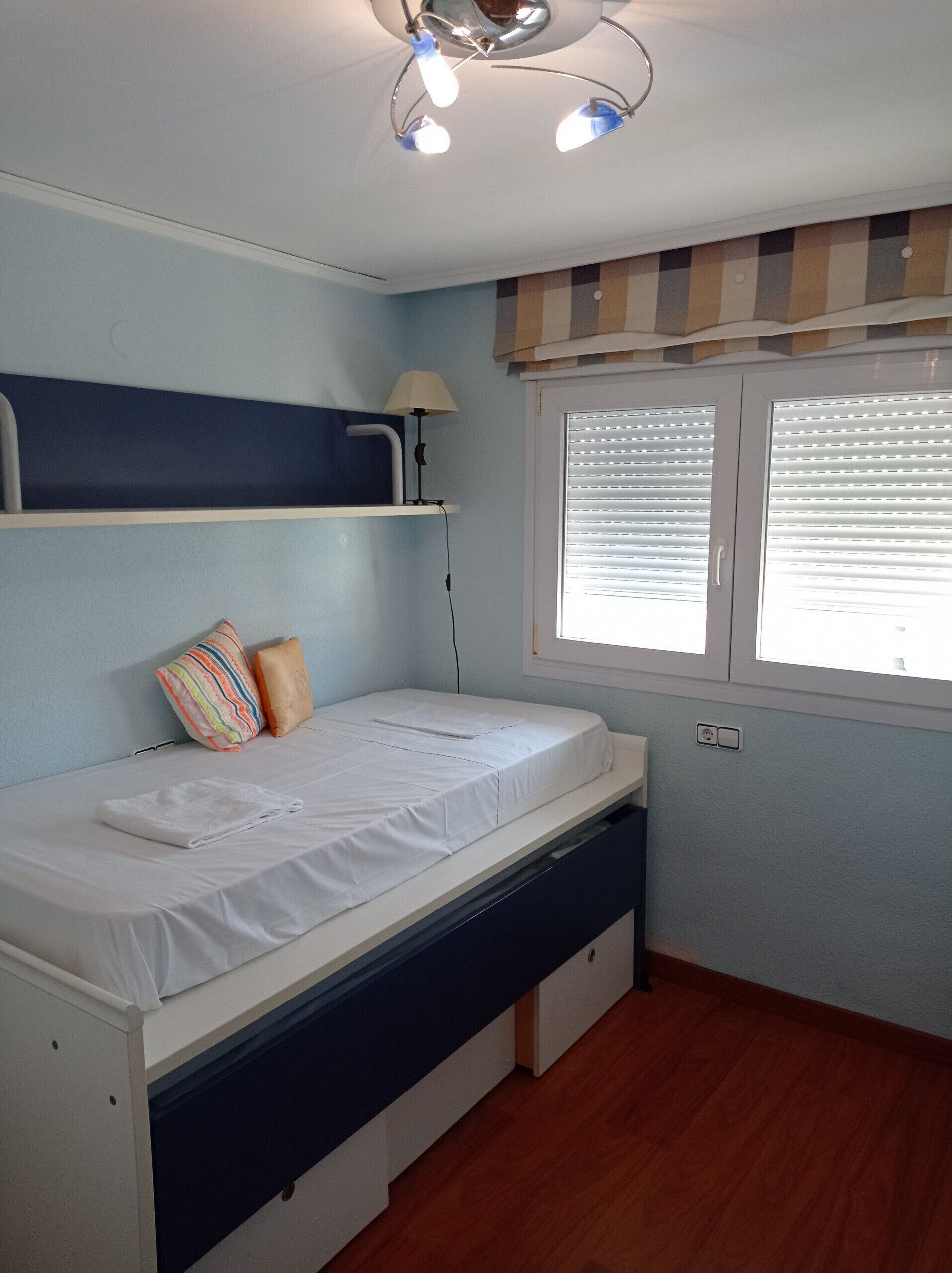 Une petite chambre à coucher, bien rangée, avec un lit simple, une table de chevet, une lampe et une fenêtre avec des stores.