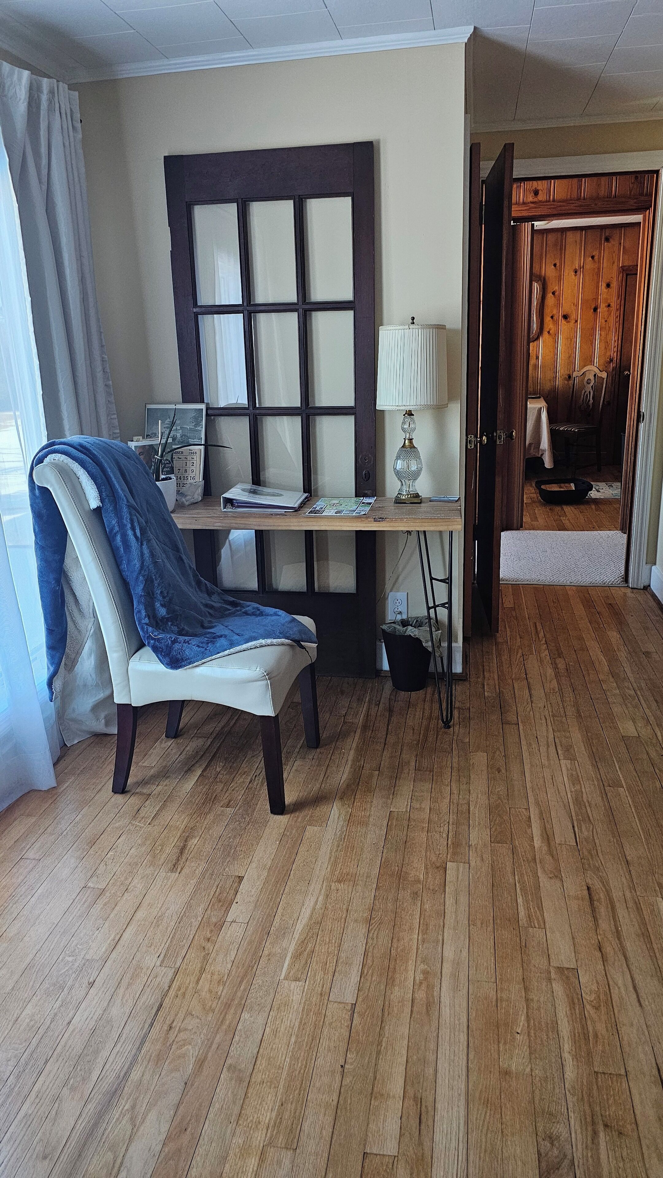 A room with a wooden floor, a desk with a lamp, a chair, and a window with curtains.