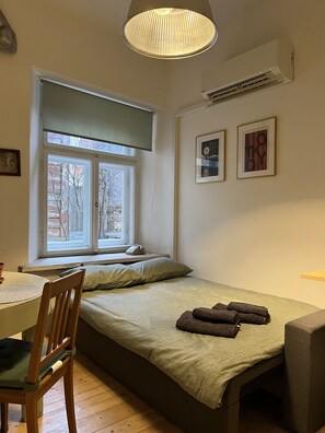 A small, cozy room with a single bed, a window with a view of trees, a table with a chair, and two framed pictures on the wall. - Old Town Apartment (Cēsis)