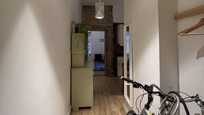 A narrow hallway with wooden flooring, a hanging light fixture, a bicycle, and a kitchen area with a refrigerator and a plant. - Old Town Apartment (Cēsis)