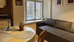 A living room with a sofa, a dining table with a plant, a clock, and a window with a view of trees. - Old Town Apartment (Cēsis)