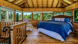 A wooden cabin bedroom with a large bed, a chair, and a window overlooking greenery. - Temple Tree Retreat: An Adventurer's Oceanfront Paradise! (Keaau)