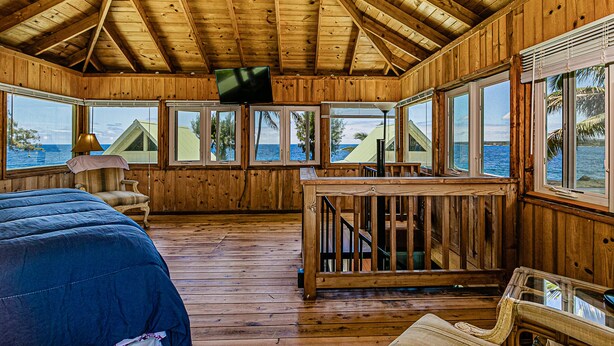 A wooden cabin with a TV mounted on the wall, large windows offering ocean views, a bed, and a small table. - Temple Tree Retreat: An Adventurer's Oceanfront Paradise! (Keaau)
