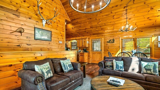 Ein Holzhütten-Interieur mit Holzwänden, einem Essbereich, zwei Ledersofas mit tierischen Kissen, einer Chandelier und einem Kamin.