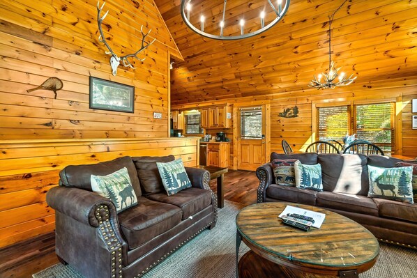 Ein Holzhütten-Interieur mit Holzwänden, einem Essbereich, zwei Ledersofas mit tierischen Kissen, einer Chandelier und einem Kamin.