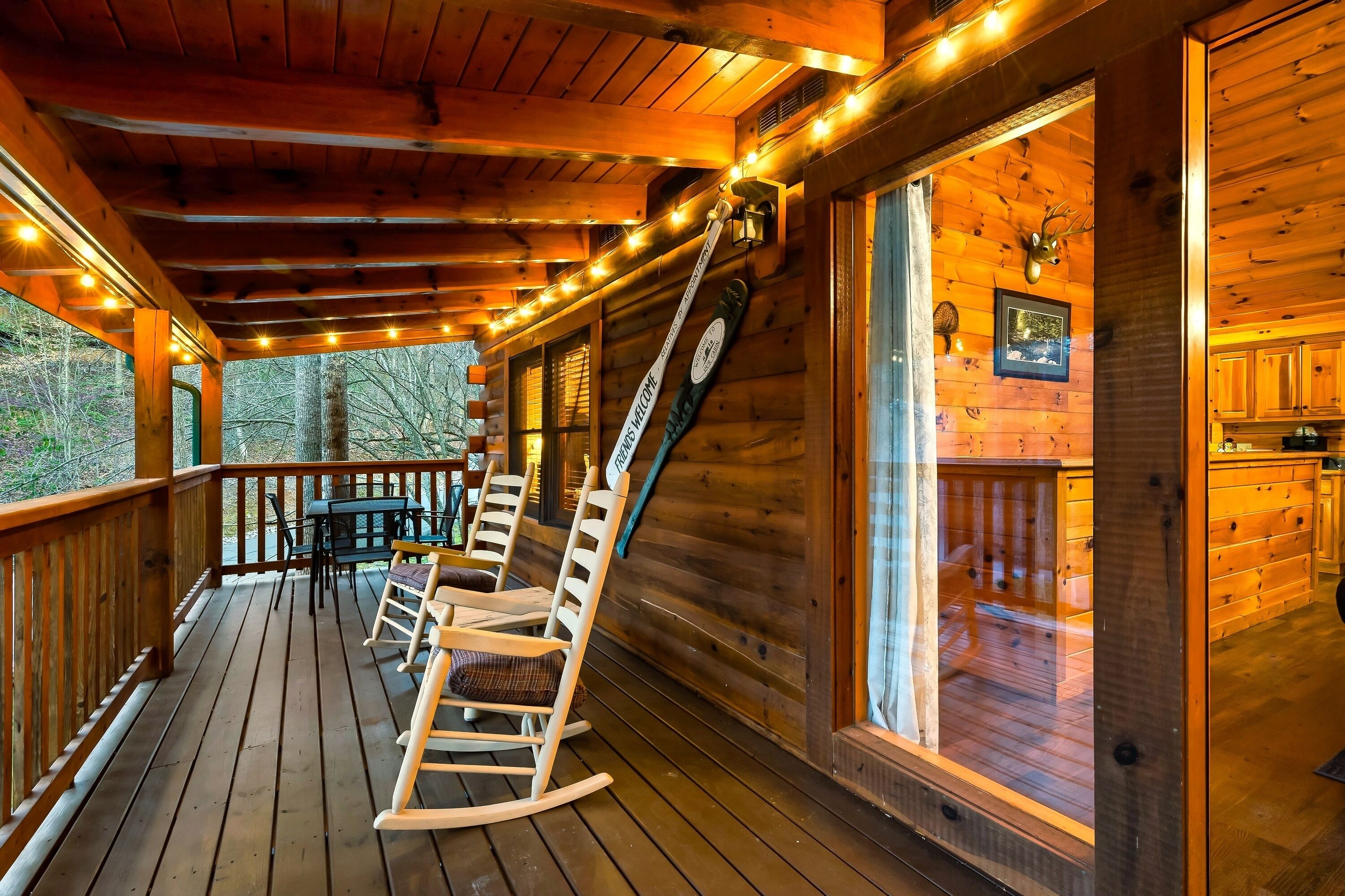 Una terraza de madera con mecedoras y una mesa, el interior de una cabaña de madera con una cocina visible a través de una ventana, y una cabeza de venado montada en la pared.