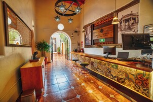 Un vestíbulo de hotel con un mostrador de recepción de madera, suelo de baldosas y un espejo grande que refleja la entrada.