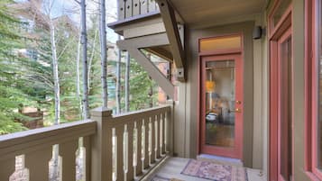 A wooden porch with a red door, a small rug, and a view of trees and buildings.