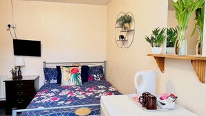 Un dormitorio con cama, mesita de noche, lámpara, televisor y estante en la pared con plantas.