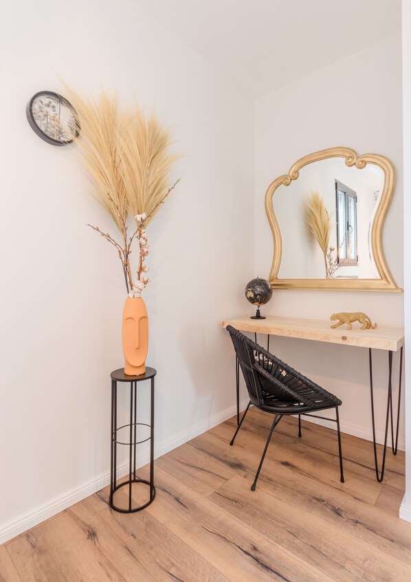 A corner room with a wooden floor, a black chair, a small round table with a vase of dried plants, a mirror with a gold frame, and a globe on the table.