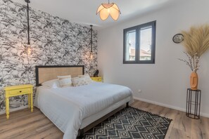 A bedroom with a bed, a yellow nightstand, a black and white patterned rug, a window, and a wall with a floral wallpaper.