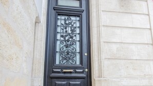 Una puerta negra con un panel de vidrio y detalles de metal decorativo, un escalón que lleva a ella y una planta en maceta a la derecha.